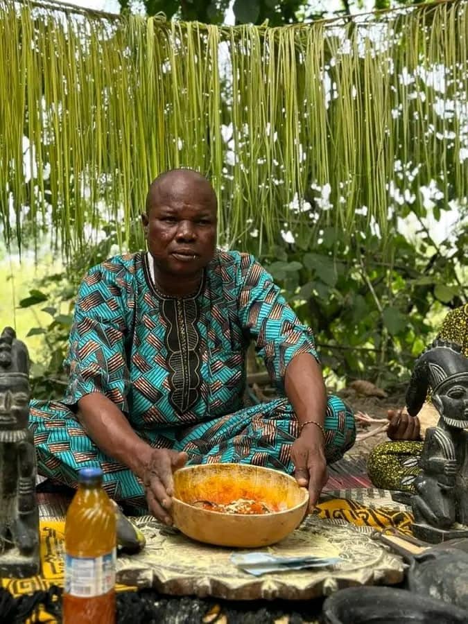 Witch Doctor Shakir in a patterned outfit, seated outdoors beside carved wooden figures